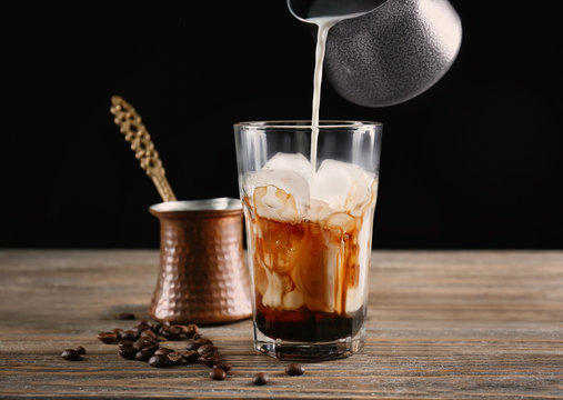Pouring milk into glass with iced coffee on black background