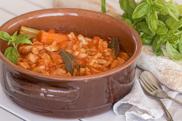 A terracotta pot with tripe with beans. A fork, a spoon, a cotton napkin, a bread and some basil.