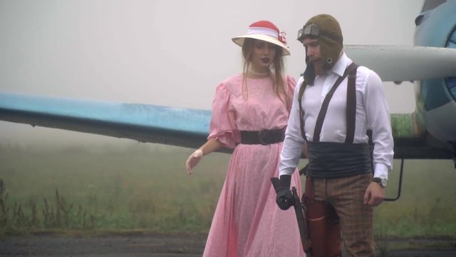 A woman in a pink dress and a white hat comes to the pilot in the helmet standing on the background of a vintage airplane, slow motion