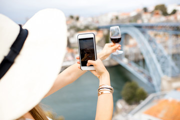 Young woman tourist photographing a glass of Porto wine sitting on the terrace with great cityscape view on Porto city in Portugal