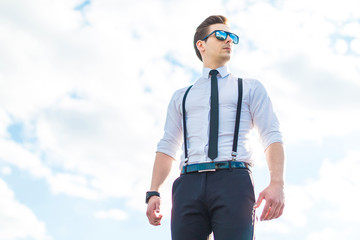 Serious young busunessman in white shirt, tie, braces and sunglasses stand on the roof