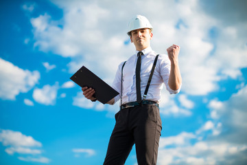 Young attractive foreman in white helmet, tie and braces stand on the roof with tablet