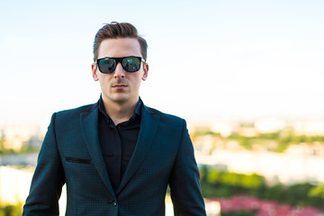 Young serious busunessman in dark suit, sunglasses, watch and black shirt stand on the roof