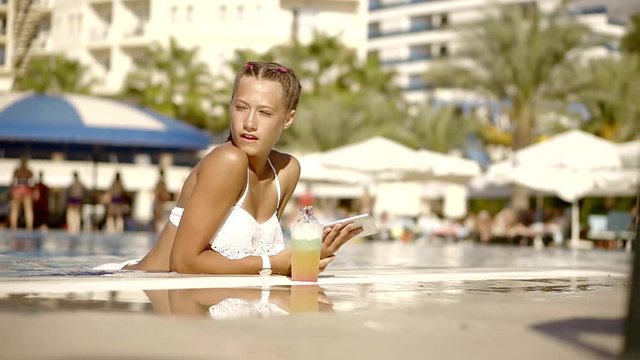 teenage girl with braids is relaxing in a swimming pool in sunny weather, holding tablet in hands