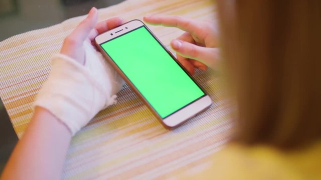 Young caucasian blonde sit at table with broken left hand and look at green screen of smartphone. Close-up view