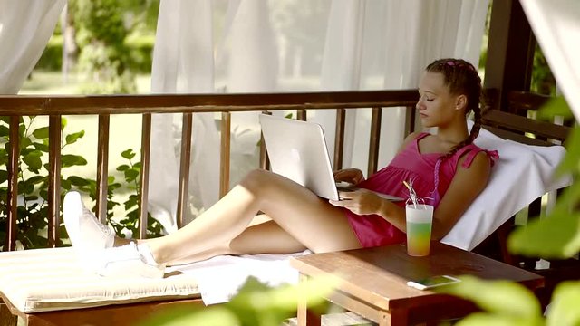 girl is lying on a sunbed under canopy in sunny weather, typing on a keyboard of a modern notebook
