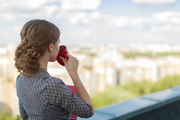 Attractive businesslady in patterned dress stand on the roof and hold paper folder and red cup