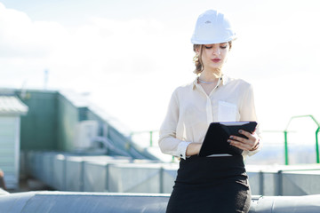 Attractive businesslady in white blouse, watch, helmet and black skirt stand on the roof and hold tablet