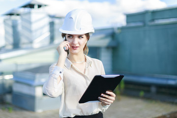 Attractive businesslady in white blouse, watch, helmet and black skirt stand on the roof, hold tablet and talk a phone