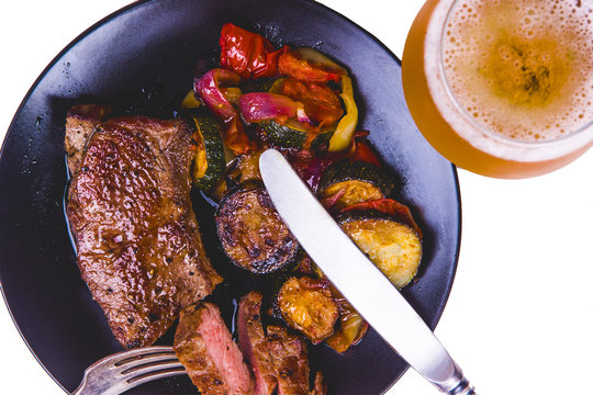 Classic New York steak with fried vegetables on a black plate and a glass of light beer - isolated