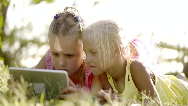 two little girlies are in sunny summer forest, lying on a grass and playing with flatbed computer