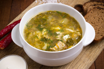 Tasty sorrel soup in a white bowl, chili pepper, sour cream, mustard beans, bread. Green borscht with beef. Wooden table. Flat lay.