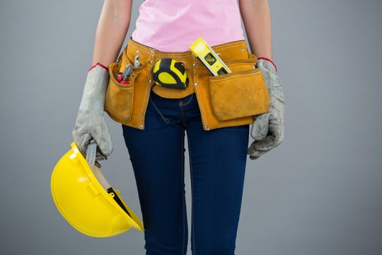 Woman With Tool Belt And Holding Hard Hat Against Grey