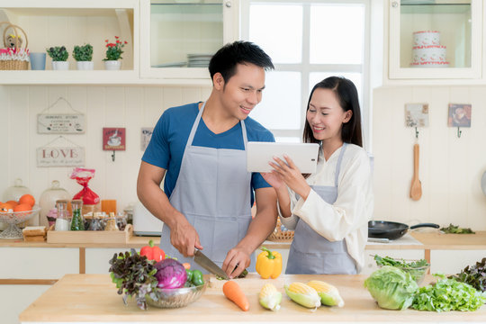 Asian beautiful young husband and wife searching menu recipe in digital tablet in kitchen at home. Happy love couple and technology concept.