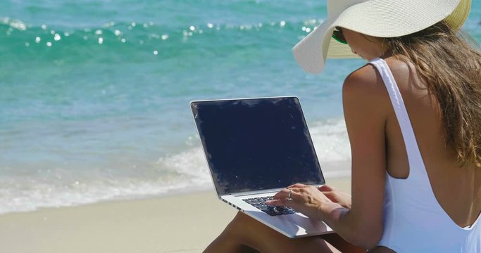 A beautiful woman on holiday with the white costume and the straw hat on the shore of a beautiful sea uses the computer to work or shop. Concept of: vacation, work, shopping,...