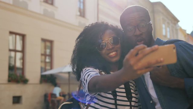 Curly girl and guy in glasses taking selfie photo with cellphone, grimacing. Nice African couple on holiday in beautiful city. Sightseeing.