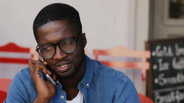 Young African man in glasses calling on mobile phone on terrace at dinner hour. Summertime. Close up.