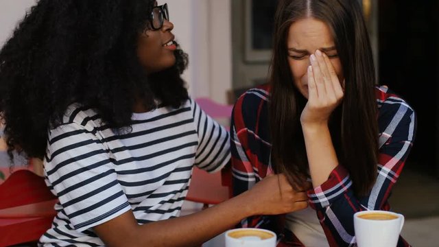 Young pretty African woman calming her disappointed friend in cafe. Outdoors. Summertime. Attractive sad girl crying.