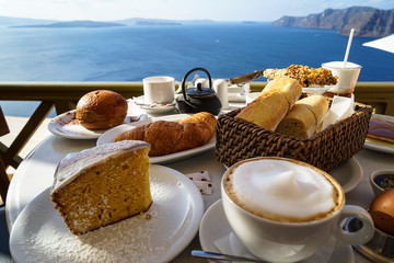 Beautiful island breakfast with Aegean sea view including cappuccino cup, cake, baguette, croissant, boiled egg, hot tea and chocolate brioche bun on white marble table