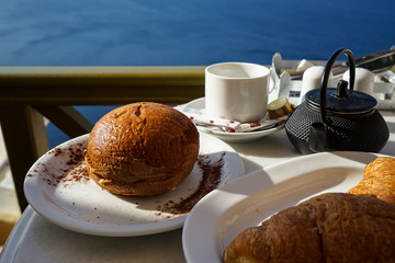 Breakfast with Aegean sea view and morning sunlight including hot tea, croissant and chocolate brioche bun on white marble table next to olive green balcony rail