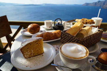Enjoy island breakfast with beautiful Aegean sea view and morning sunlight including cappuccino cup, cake, baguette, croissant, boiled egg, hot tea and chocolate brioche bun on white marble table