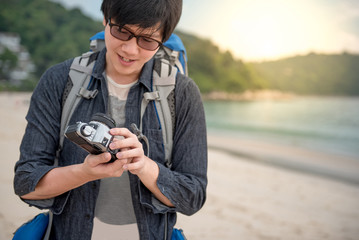 Young Asian man traveler look at his vintage camera, Happy male photographer on the beach, summer holiday and vacation lifestyle concepts