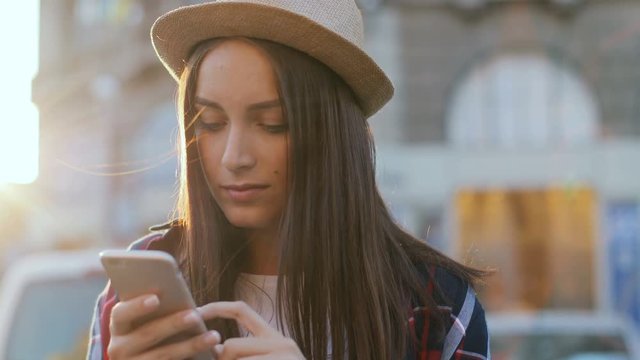 Attractive young modern woman in hat sitting on bench on sunny day. Girl holding smartphone in hands and reading text messages in street. Outdoors.