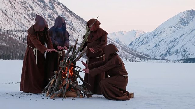 Group of monks in brown cloaks of the order of St. Francis are warming themselves by the fire
