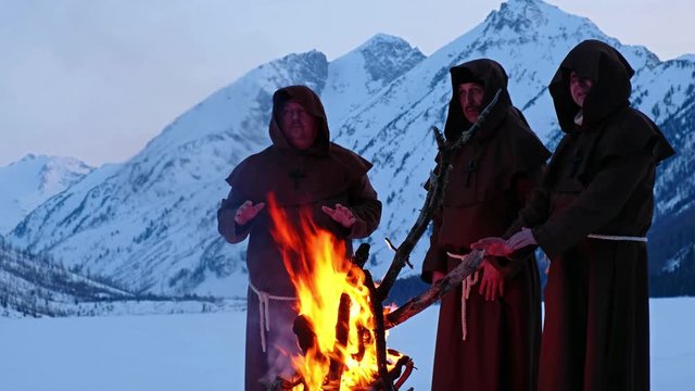 Group of monks in brown cloaks of the order of St. Francis are warming themselves by the fire