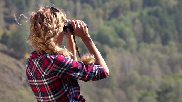 Young blonde woman tourist   on a cliff looking through binoculars on the landscape with the river