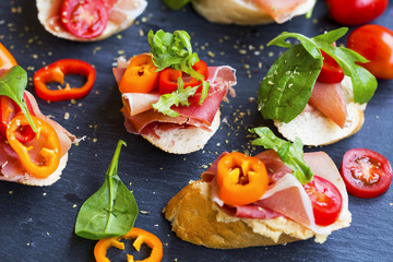 Canapes appetizer with prosciutto ham, pepper slices, tomatoes and green spinach and rucola leaves, healthy crostini or bruschettas italian appetizer seasoned