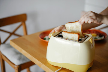 yellow toaster with toasted bread for breakfast inside, on the table in the kitchen interior. Hands Girl pulls out ready toasts. - obrazy, fototapety, plakaty