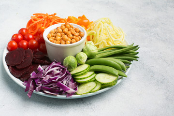 Vegan eating concept, healthy salad bowl, colourful vegetables, carrot, courgette, cabbage, chickpeas, cucumber and tomatoes, on wooden board on white table, copy space, top view, selective focus