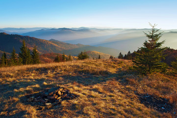 Spectacular view of hills of smoky mountain range covered in purple grey mist; abandoned stones of campsite fireplace under blue cloudless sky on a warm fall evening in October. Carpathians, Ukraine