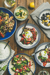 Flat-lay of healthy dinner table setting. Fresh salad, grilled vegetables with yogurt sauce, pickled olives, lemon water over wooden background, top view. Clean eating, vegan, vegetarian food concept