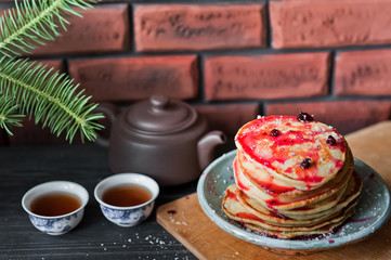 Stack of fritters with blueberries. Pancakes with red jam and coconut shavings. A small teapot with tea bowls. Cashew and coconut chips on a dark background. Sprig of Christmas tree on the table.