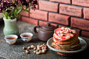 Stack of fritters with blueberries. Pancakes with red jam and coconut shavings. A small teapot with tea bowls. Cashew and coconut chips on a dark background. Sprig of Christmas tree on the table.