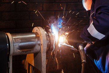 Close-up male auto mechanic in blue uniforms cooking metal on an automatic welding machine for the repair of cardan shafts in an industrial workshop, from the welding apparatus smoke rises