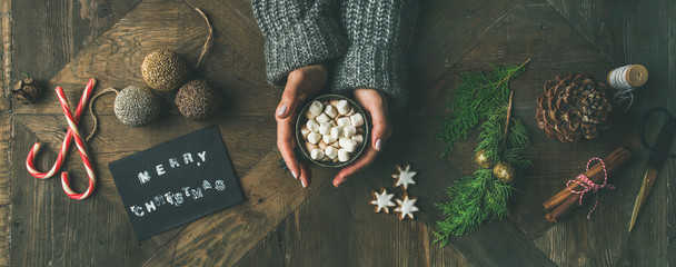 Christmas, New Year background. Flat-lay of greeting card, glittering toys, woman's hands in grey knitted sweater holding mug of hot chocolate, cinnamon, scissors over wooden table, top view