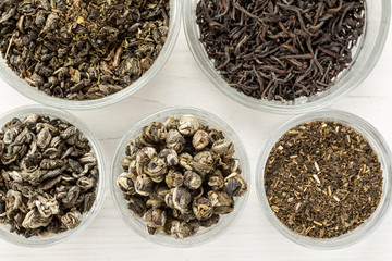 Different dry tea in a small glass dishes on a white wooden table, top view