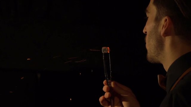 Back close-up view of the man blows on the piece of hookah coal in the dark room. The view of falling sparkls.