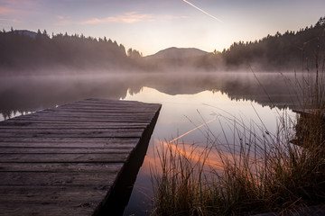 Morgendämmerung am See mit Gräser und Steg im Vordergrund und Nebel im Hintergrund