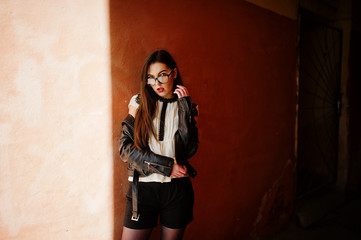 Stylish brunette girl wear on leather jacket and shorts with glasses against orange wall at shadows.