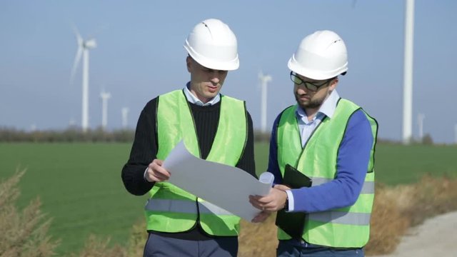 Two engineers discussing against turbines on wind farm on sunny day