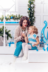 beautiful mother and charming daughter, sitting next to a Christmas tree and Christmas balls in warm stylish sweater and jeans