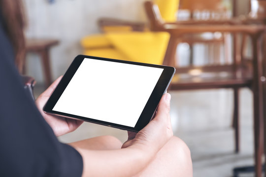 Mockup image of woman's hands holding black tablet pc with blank white screen on thigh with concrete floor background in modern cafe
