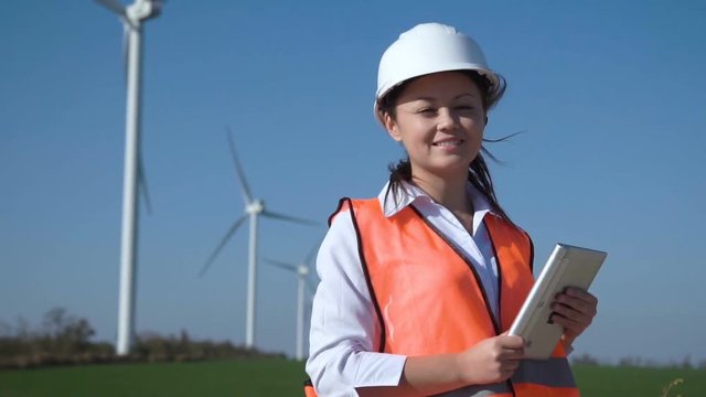 Slow motion of cheerful korean woman with digital tablet wearing hard hat standing against turbines at wind farm on sunny day