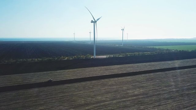 Aerial shot of the rotating wind turbines in the field with the tractor which cultivates the soil