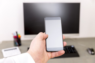 Closeup of a man holding smartphone with a blank screen monitor in the hands, working behind a computer keyboard on wooden table in the office 