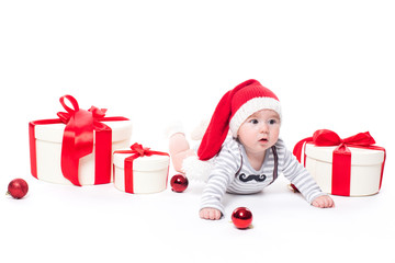 Cute baby in a red New Year's cap with a smile on his face lying on his stomach on a white background surrounded by boxes with gifts, and blue and red Christmas balls